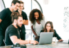 A diverse group of young professionals gathered around a laptop, collaborating on a digital marketing project in a bright, modern office. 