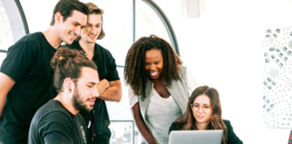 A diverse group of young professionals gathered around a laptop, collaborating on a digital marketing project in a bright, modern office. 